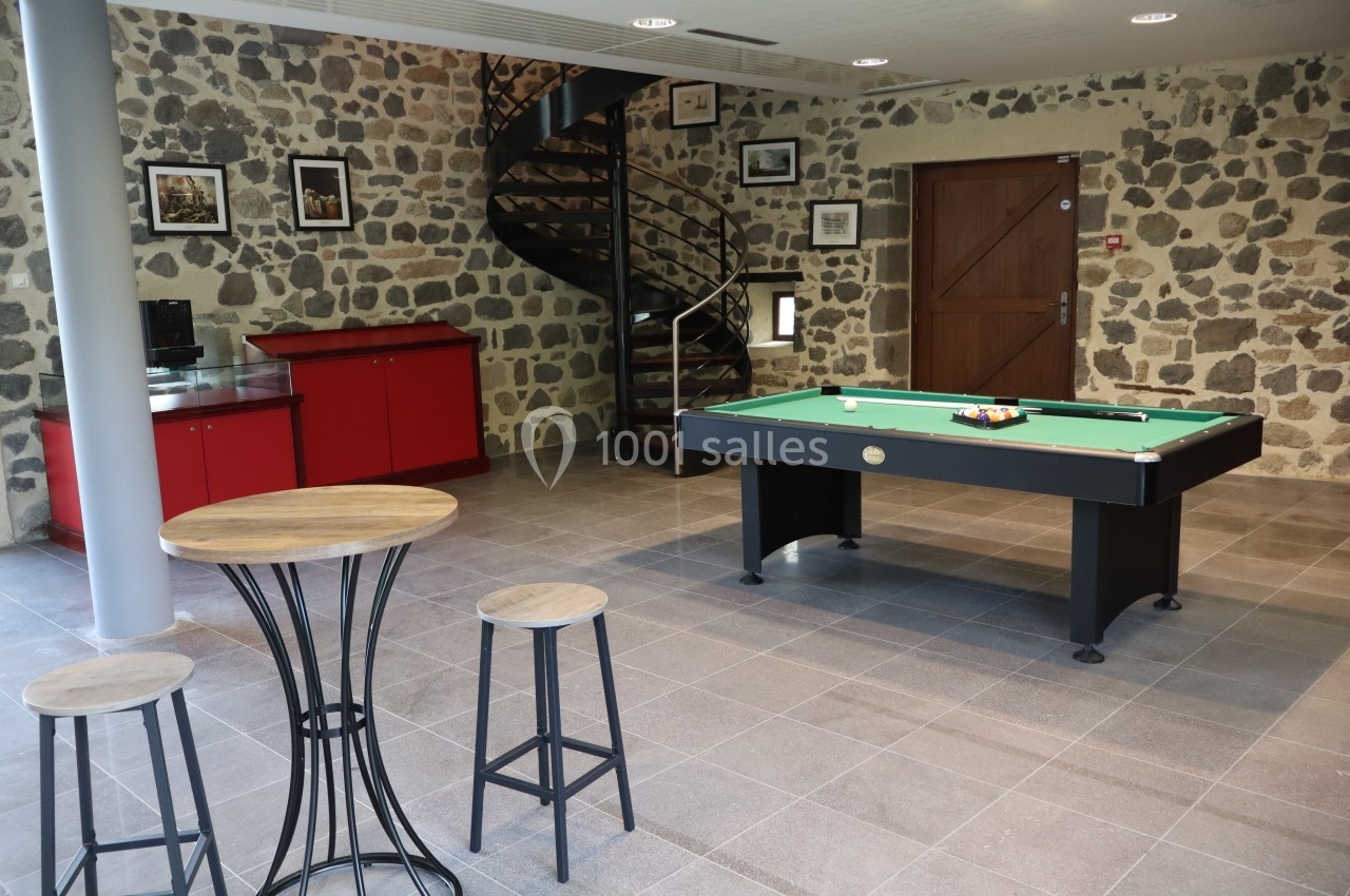Salle avec un billard, un bar rouge, des tabourets en bois et un escalier en colimaçon sur fond de mur en pierre.