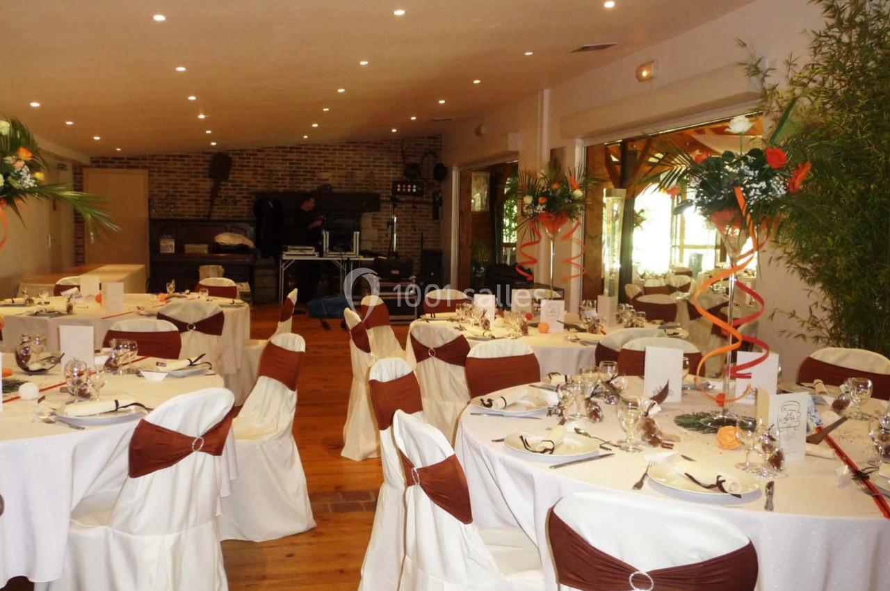 Salle de réception décorée avec des tables dressées, nappes blanches, chaises ornées de nœuds marron et arrangements floraux.
