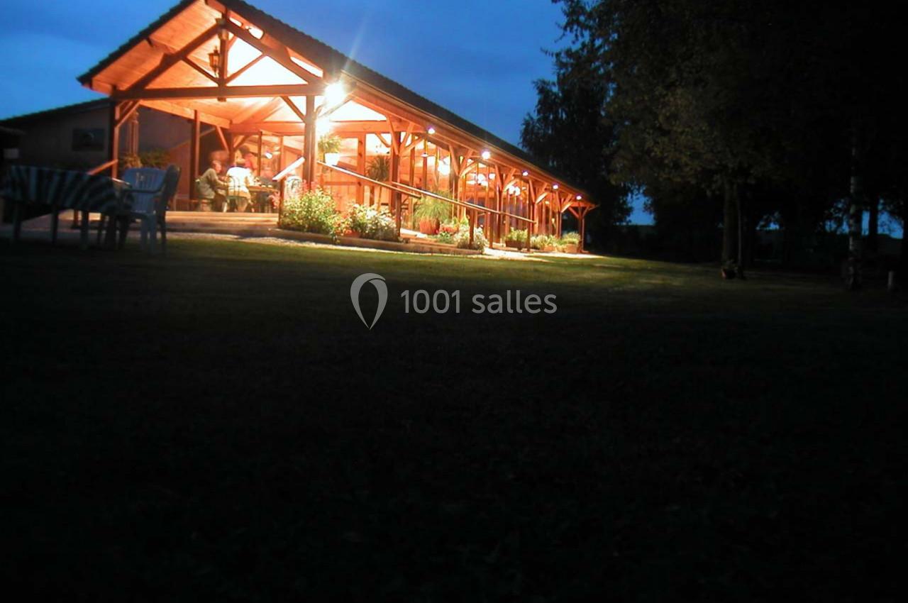 Terrasse en bois éclairée le soir, entourée de végétation, avec des personnes assises à des tables.