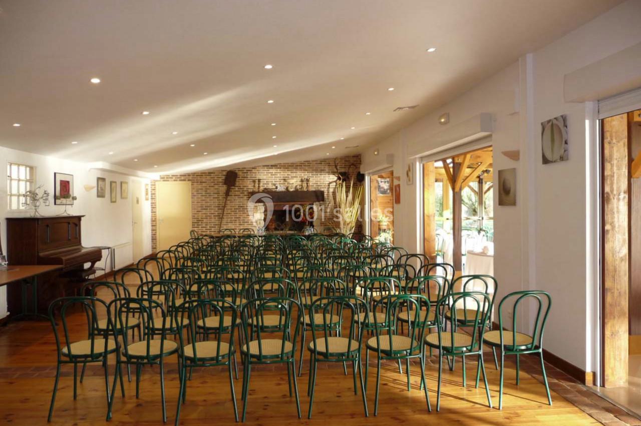 Salle lumineuse avec des rangées de chaises vertes en bois, un piano à gauche et une cheminée en briques au fond.