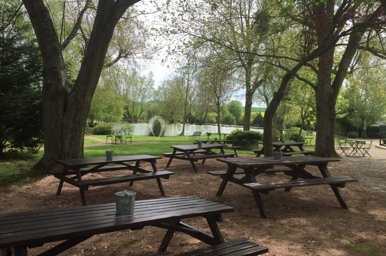 Tables de pique-nique en bois disposées sous des arbres près d'un étang dans un cadre naturel verdoyant.