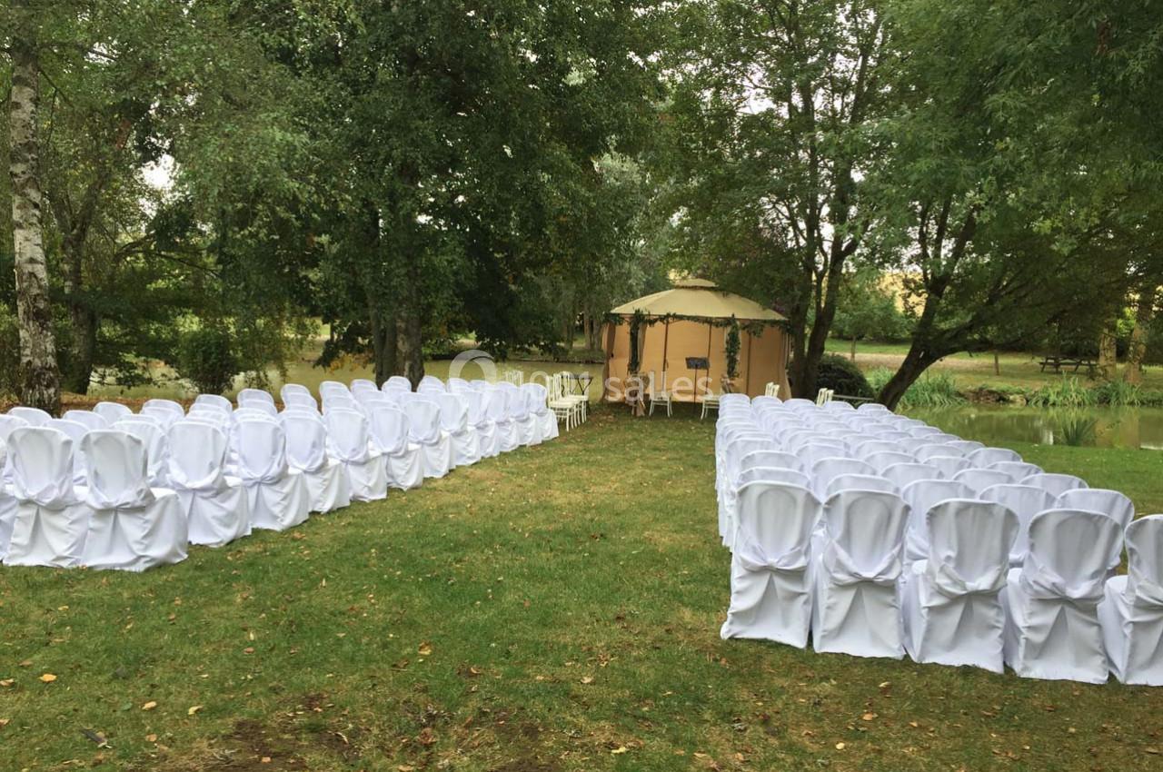Chaises blanches disposées en deux rangées sur une pelouse, face à une tente beige entourée d'arbres.