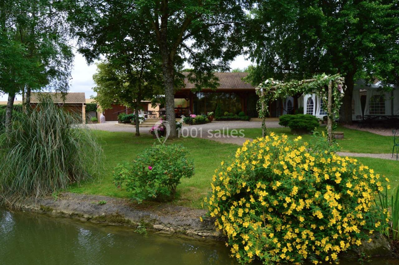 Jardin verdoyant avec des fleurs jaunes, arbres et un étang au premier plan, maison et pergola en arrière-plan.