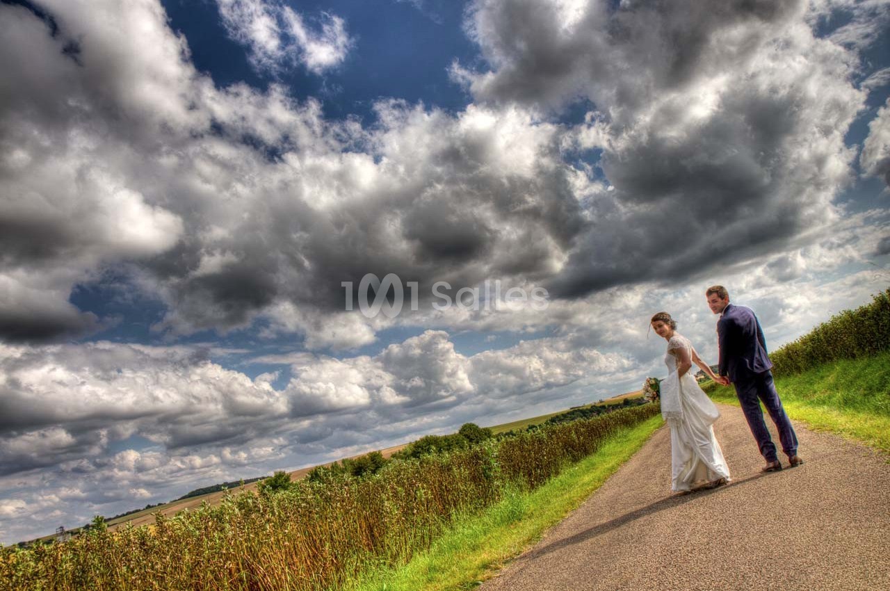 Un couple en tenue de mariage marche sur une route de campagne sous un ciel nuageux.