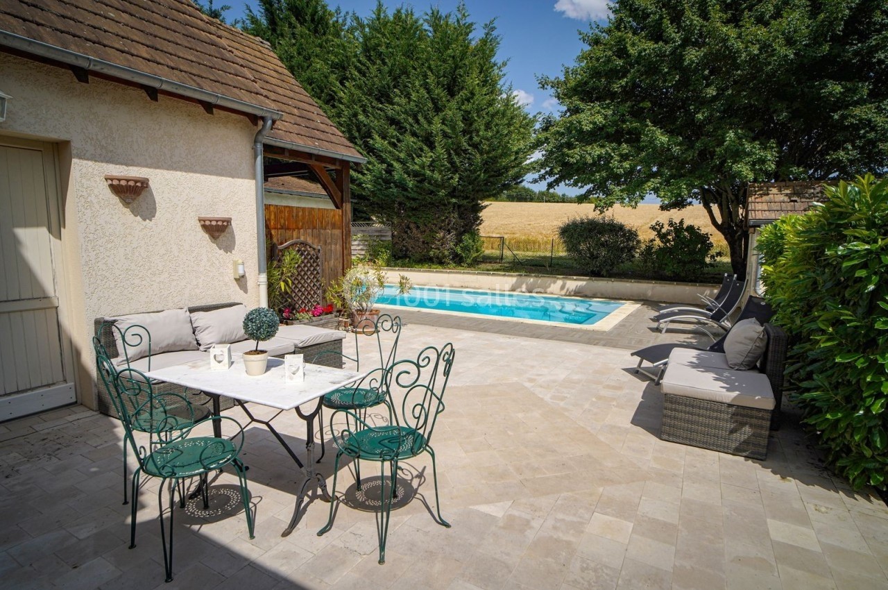 Terrasse avec table et chaises en fer forgé, piscine entourée de transats, arbres et champs en arrière-plan.