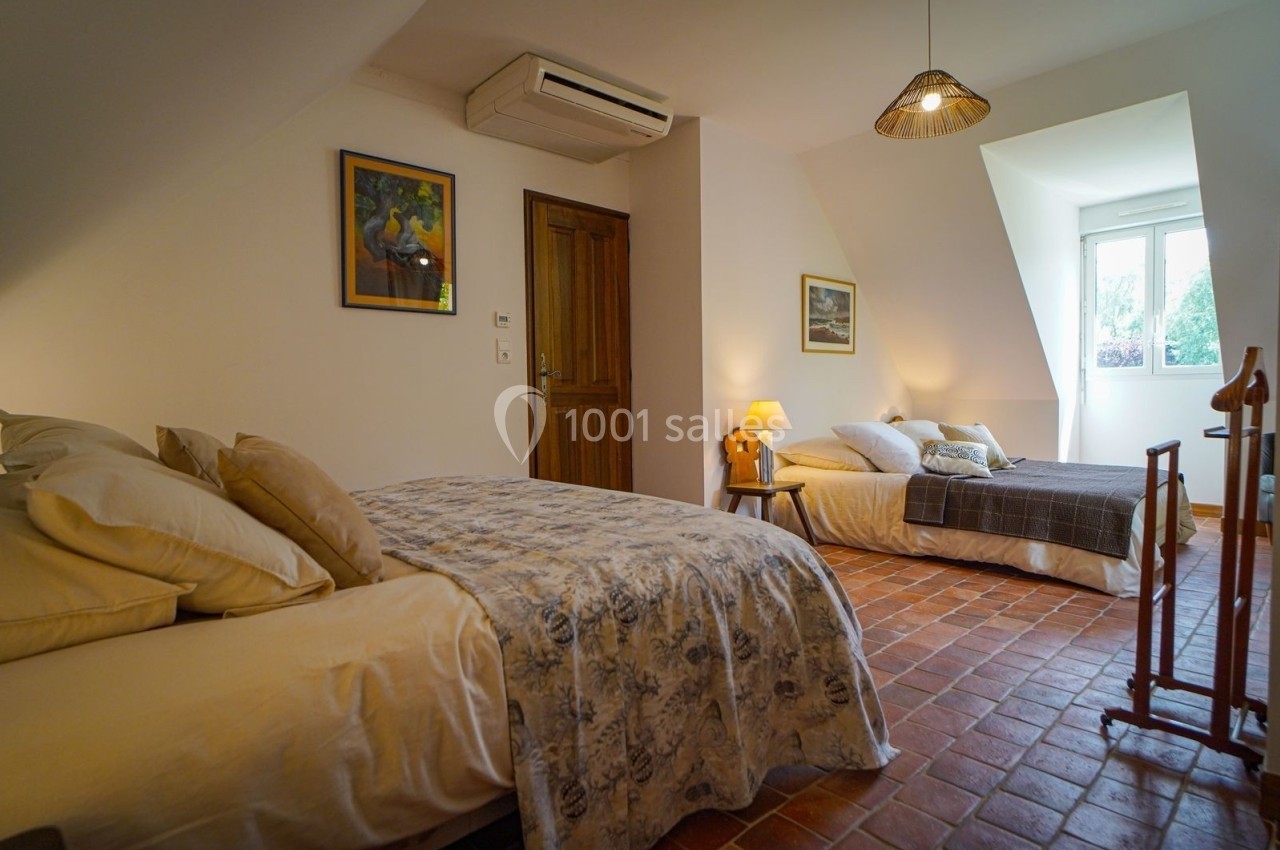 Deux lits dans une chambre lumineuse avec sol en terre cuite, murs blancs, tableaux et fenêtre donnant sur l'extérieur.