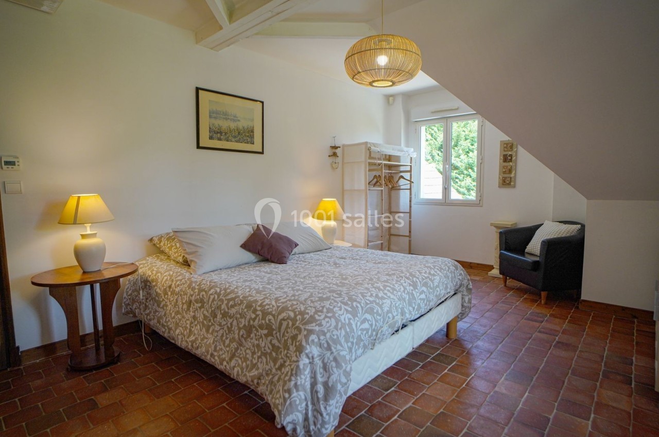 Chambre lumineuse avec lit double, lampes de chevet, fauteuil, fenêtre donnant sur un espace vert et sol en terre cuite.