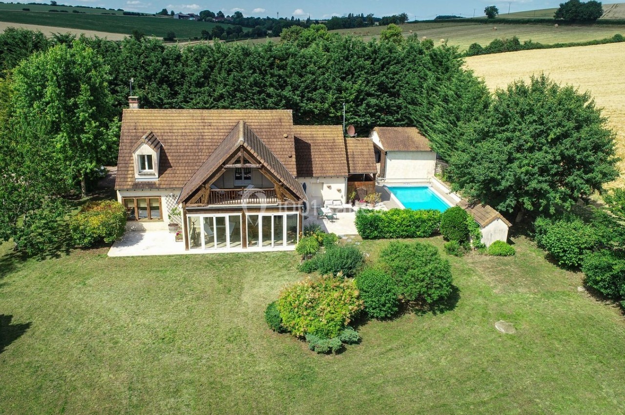 Vue aérienne d'une maison avec toit en pente, jardin arboré, piscine et champs environnants.