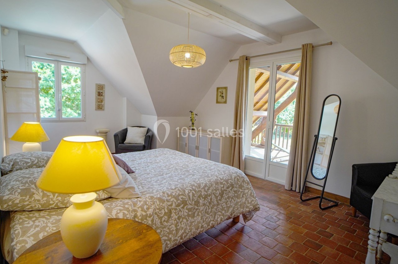 Chambre lumineuse avec lit double, lampes de chevet, sol en terre cuite, miroir sur pied et accès à un balcon en bois.