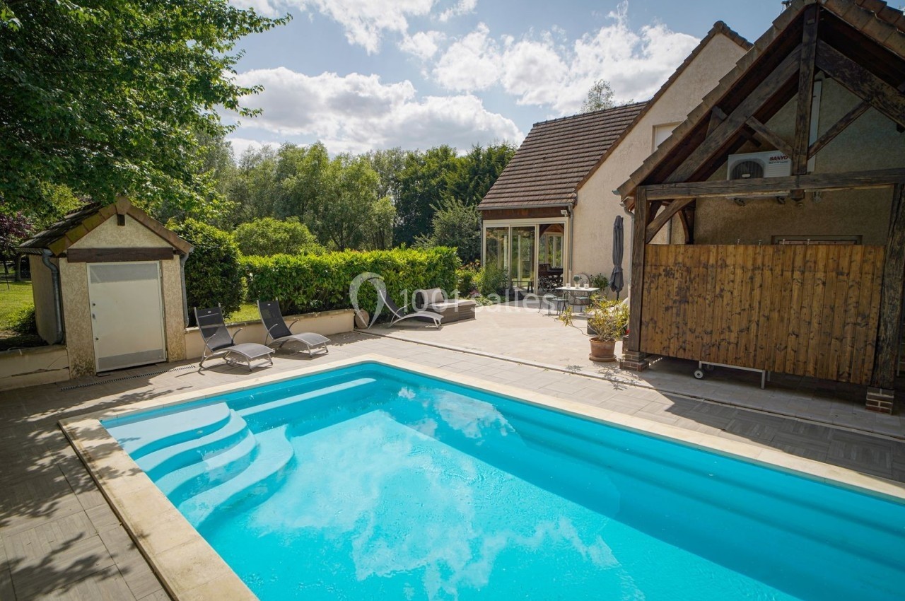 Piscine extérieure entourée de chaises longues, terrasse en bois et maison avec jardin arboré en arrière-plan.
