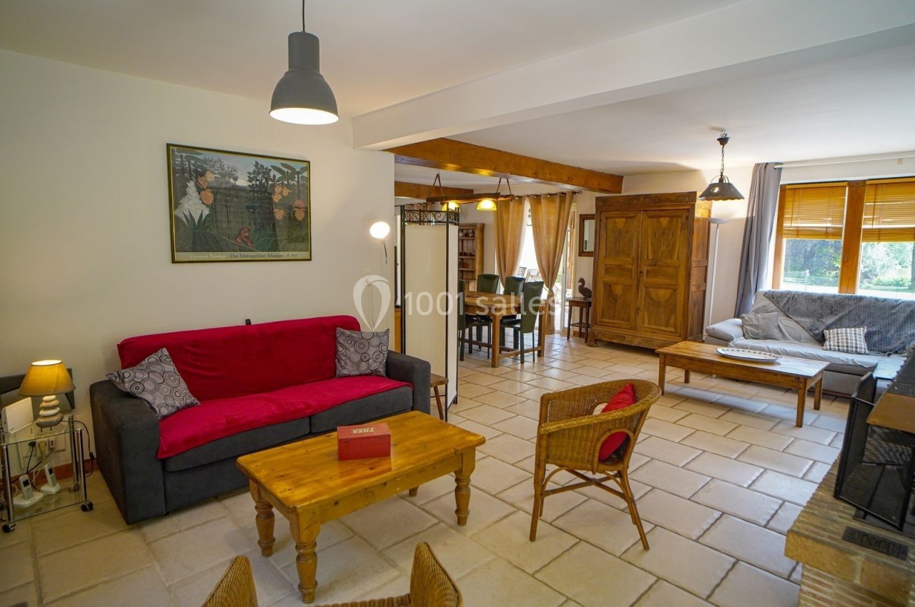 Salon lumineux avec canapé rouge, fauteuils en osier, table basse en bois et vue sur une salle à manger en arrière-plan.