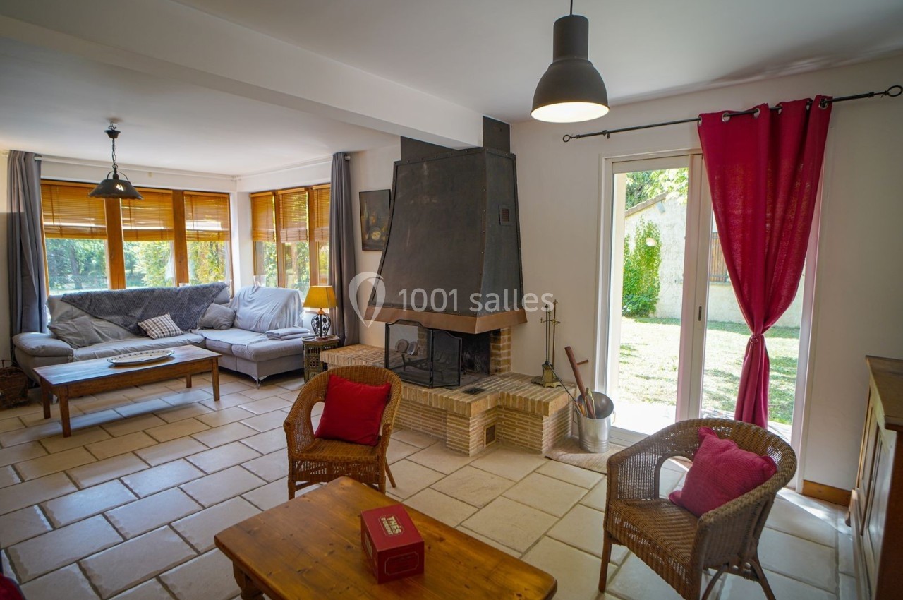 Salon lumineux avec cheminée, canapé d'angle, fauteuils en osier, table basse en bois et vue sur un jardin verdoyant.