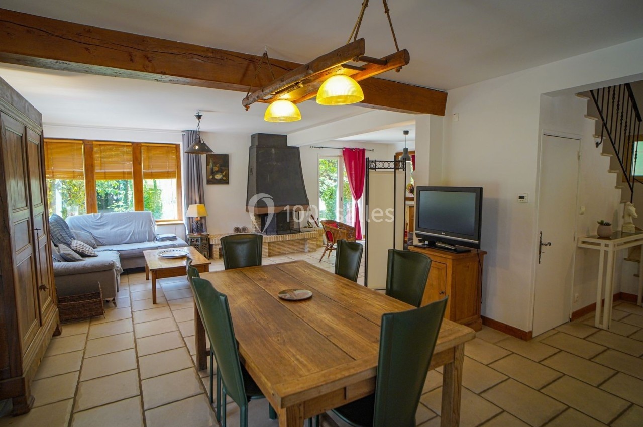 Salle à manger lumineuse avec table en bois, chaises vertes, cheminée, canapé et télévision dans un intérieur chaleureux.