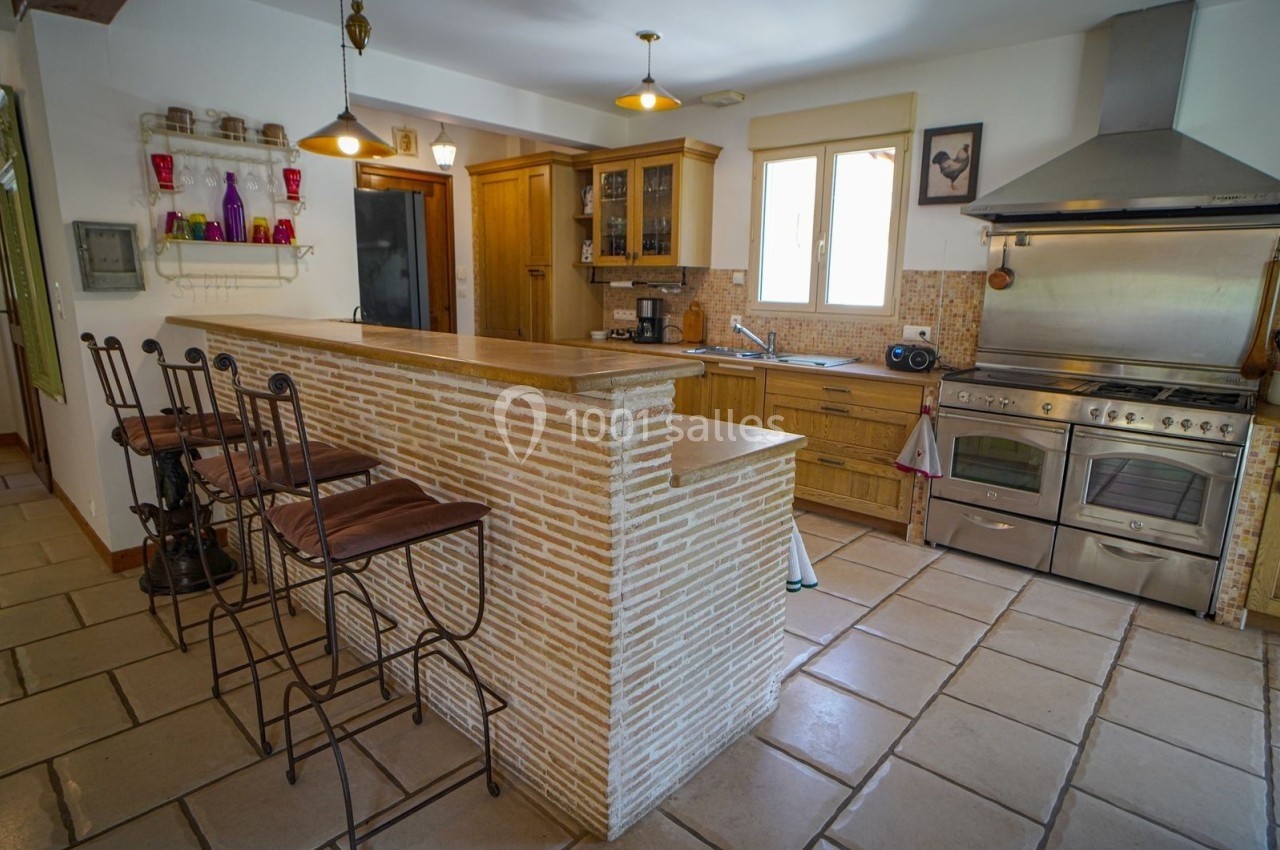 Cuisine équipée avec un îlot central en briques, tabourets hauts, cuisinière en inox et rangements en bois clair.