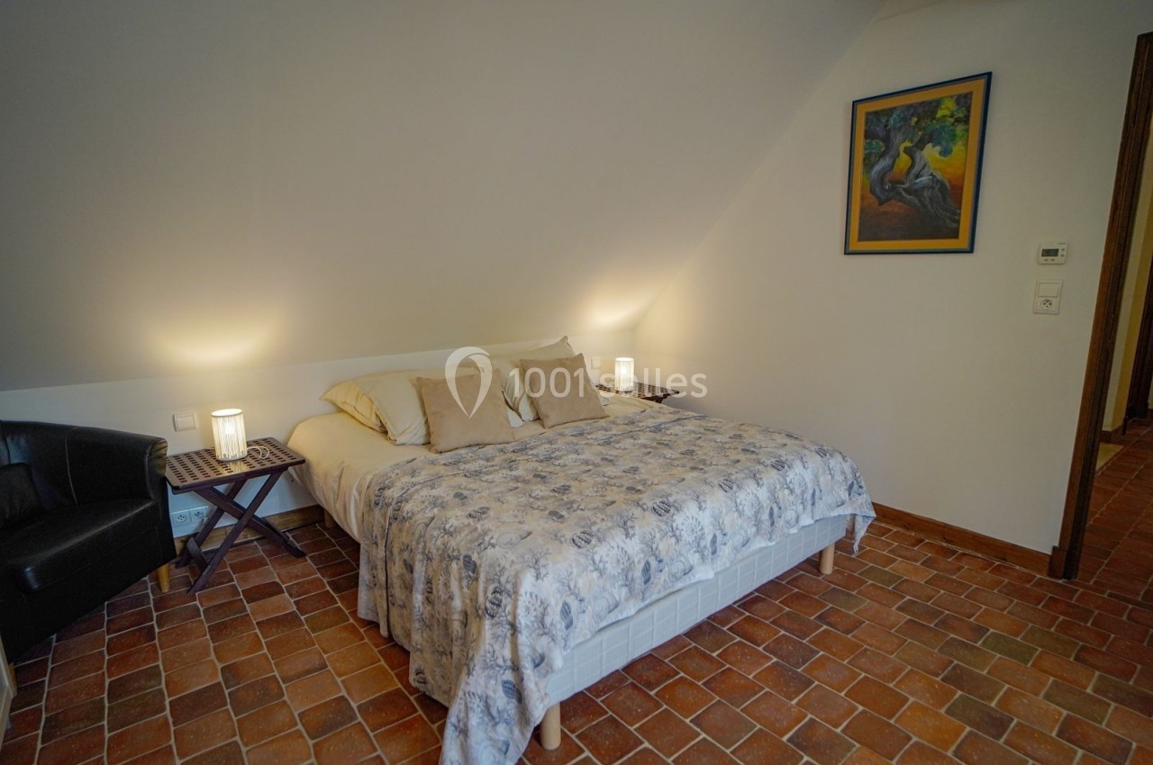Chambre avec lit double, couvre-lit fleuri, sol en terre cuite, fauteuil noir et éclairage doux.