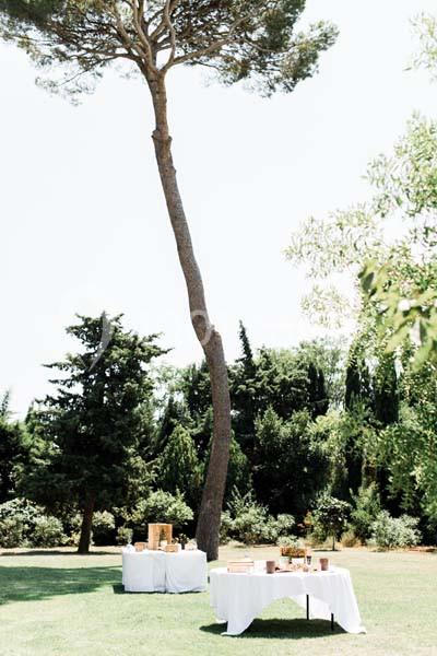 Location salle Pomérols (Hérault) - Domaine Fon de Rey #10 Deux tables dressées avec nappes blanches dans un jardin verdoyant, près d'un grand pin élancé.
