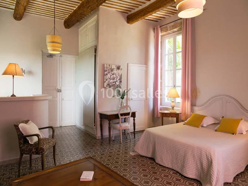 Location salle Pomérols (Hérault) - Domaine Fon de Rey #15 Chambre lumineuse avec lit double, murs clairs, poutres apparentes, mobilier en bois et grande fenêtre avec rideaux roses.