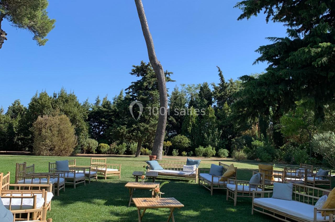 Location salle Pomérols (Hérault) - Domaine Fon de Rey #44 Chaises et canapés en bois disposés sur une pelouse verdoyante entourée d'arbres sous un ciel bleu.