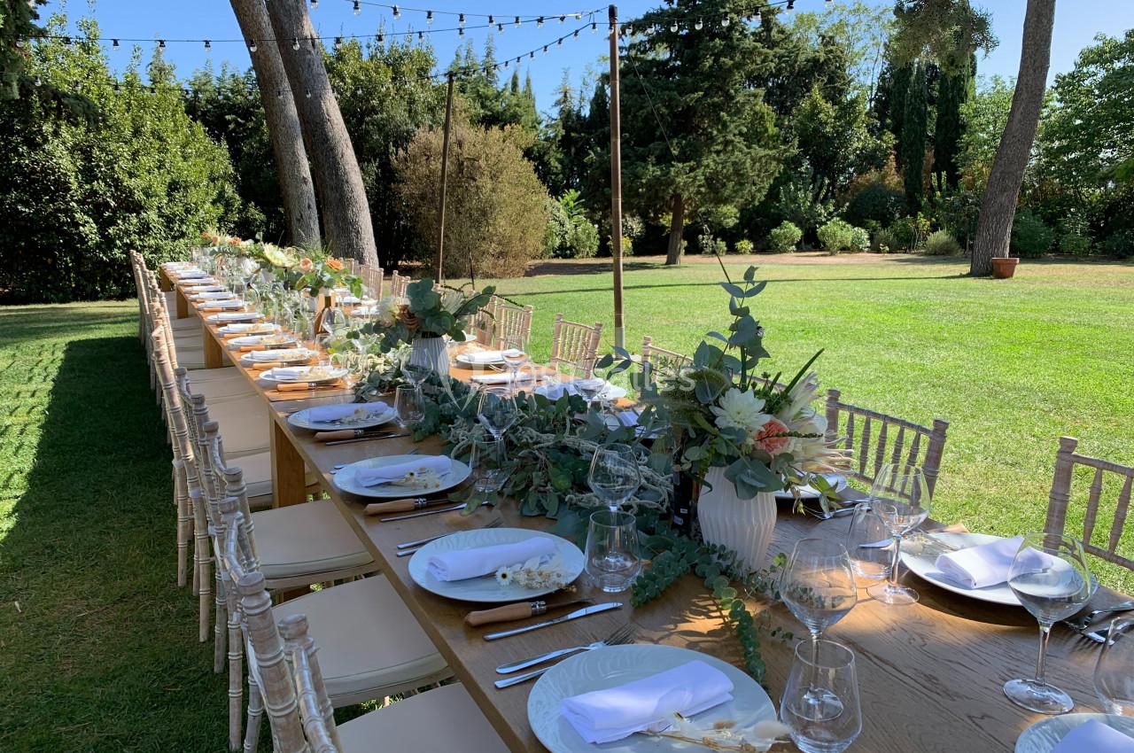 Location salle Pomérols (Hérault) - Domaine Fon de Rey #46 Grande table en bois décorée de fleurs et vaisselle élégante, installée dans un jardin sous des guirlandes lumineuses.
