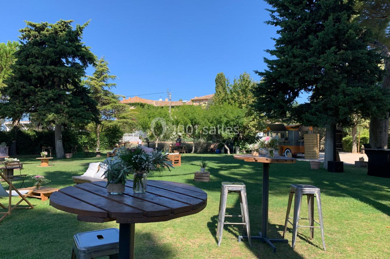 Location salle Pomérols (Hérault) - Domaine Fon de Rey #56 Espace extérieur avec tables hautes, tabourets et décorations florales, entouré d'arbres et de verdure sous un ciel bleu.