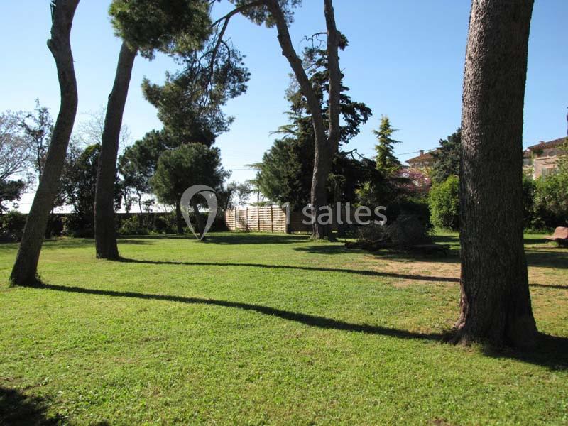 Location salle Pomérols (Hérault) - Domaine Fon de Rey #20 Pelouse verdoyante avec des arbres dispersés sous un ciel bleu clair, entourée de végétation et de clôtures en bois.