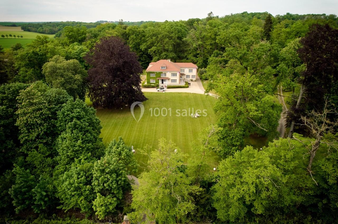 Vue aérienne d'une maison entourée d'un grand jardin verdoyant et d'une forêt dense en arrière-plan.