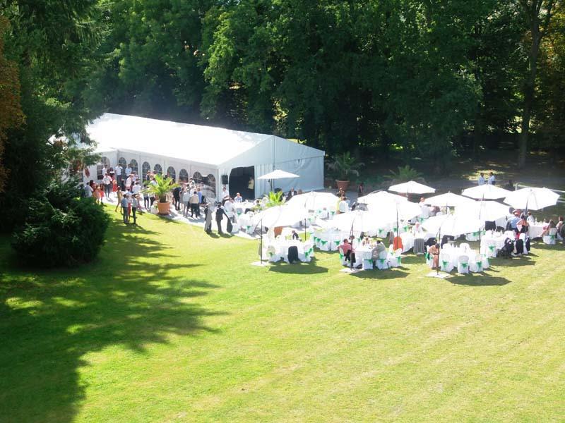 Vue aérienne d'une réception en plein air avec une tente blanche, des tables sous des parasols et des invités dans un parc…