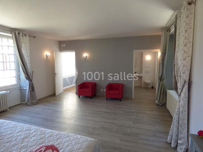 Chambre spacieuse avec parquet, deux fauteuils rouges, lit, grandes fenêtres et accès à une salle de bain attenante.