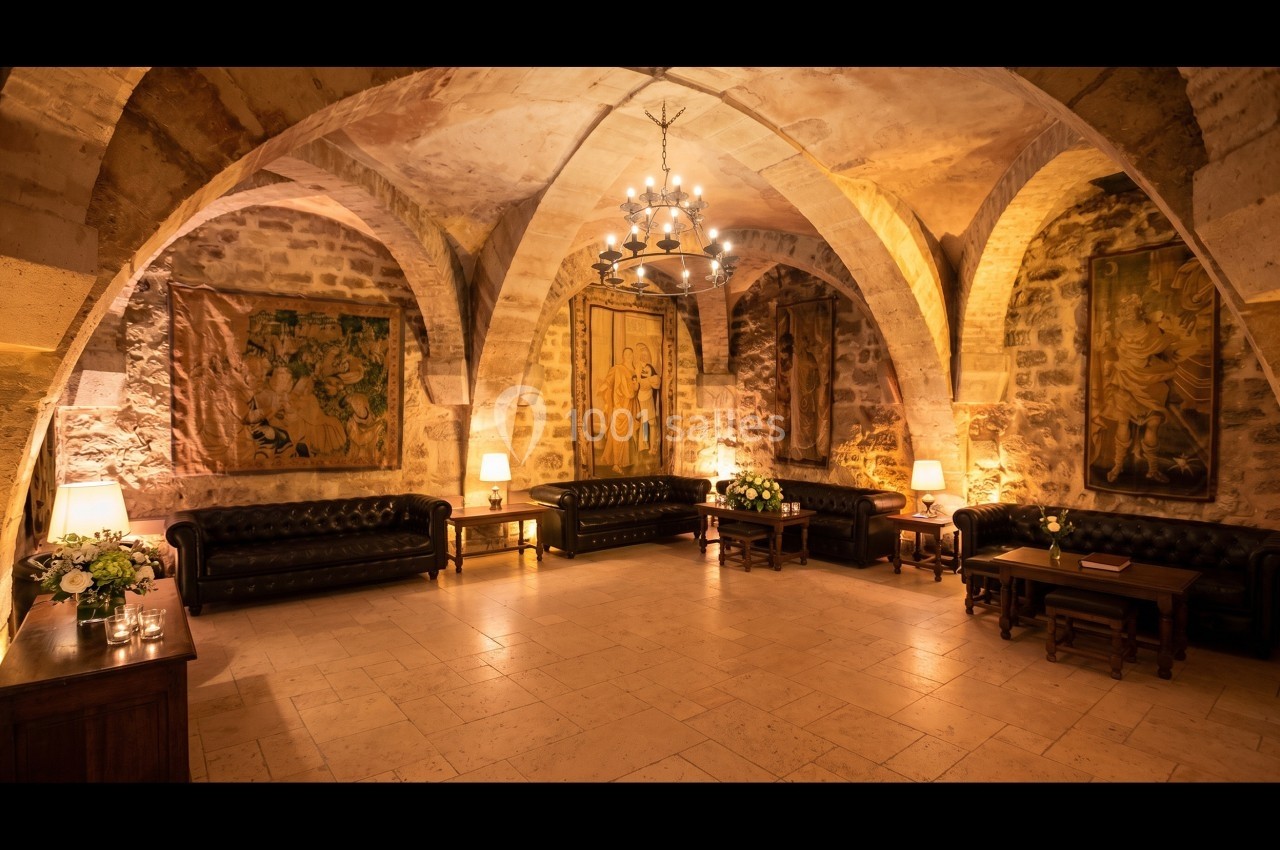 Salle voûtée en pierre avec canapés en cuir, tapisseries murales, tables en bois et éclairage chaleureux.