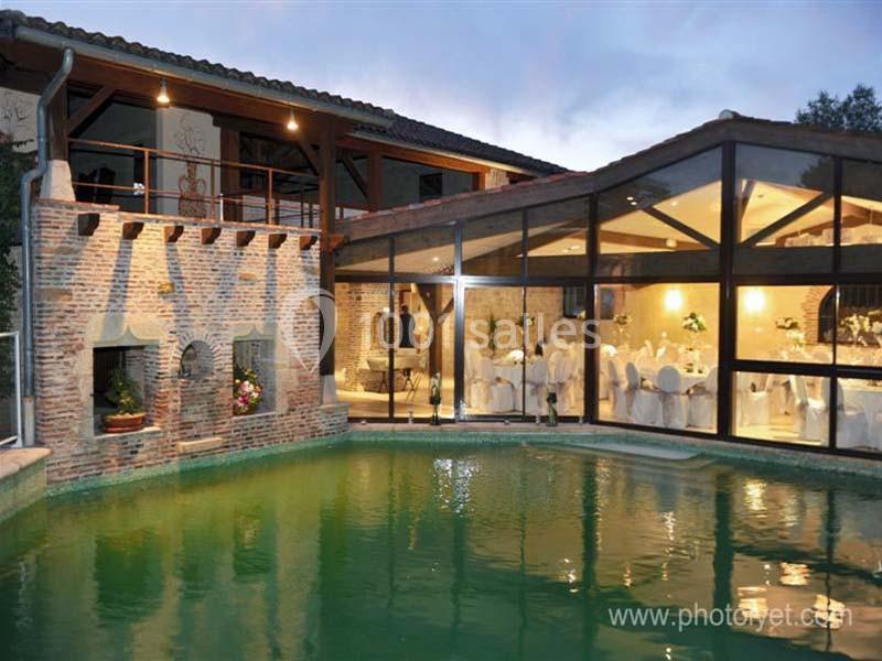 Vue d'une bâtisse en pierre avec une piscine éclairée et une salle de réception vitrée en arrière-plan.