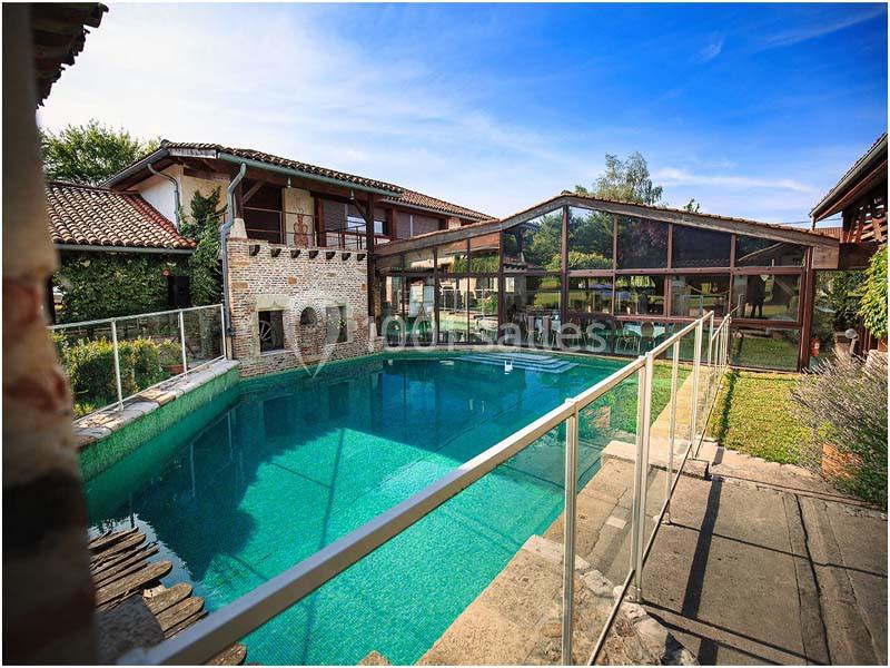 Piscine extérieure entourée de bâtiments en pierre et en bois, avec une barrière en verre et un ciel dégagé.