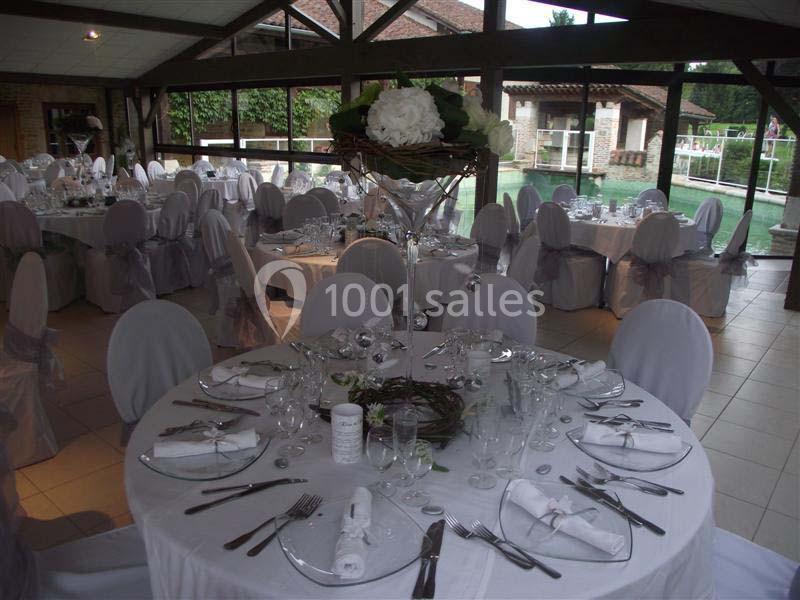 Salle de réception avec tables rondes dressées, nappes blanches, chaises couvertes et décoration florale centrale.