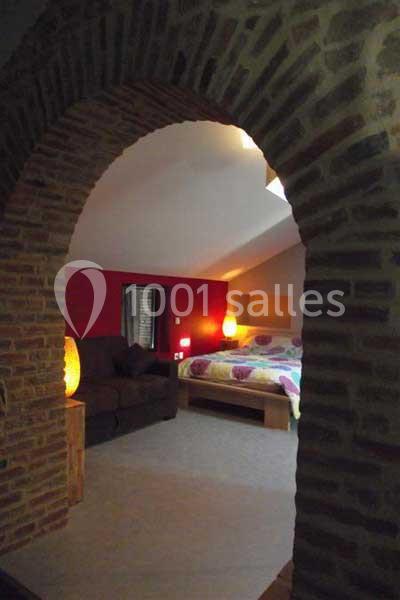 Vue d'une chambre avec un lit double, un canapé, des lampes allumées et un mur rouge, encadrée par une arche en briques.