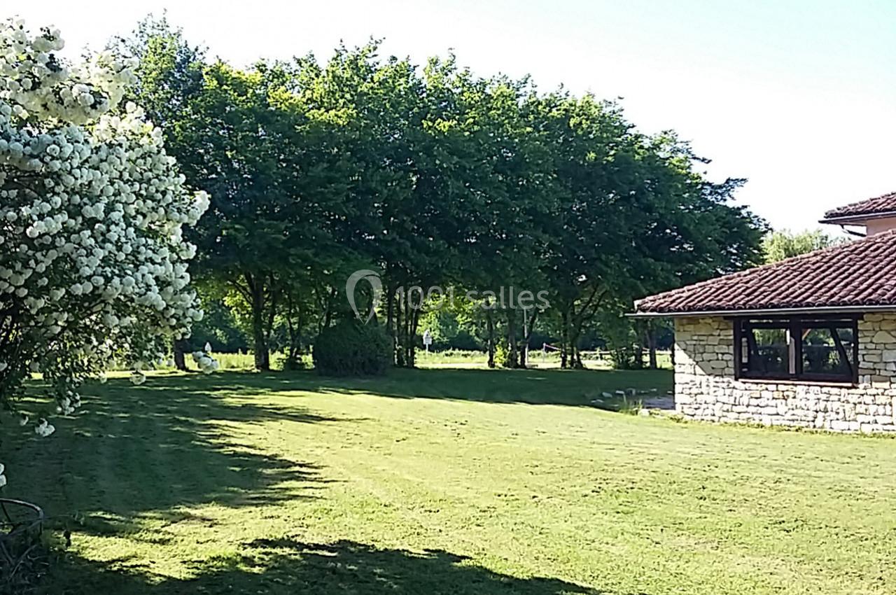 Jardin verdoyant avec pelouse, arbres en arrière-plan et maison en pierre à droite sous un ciel dégagé.