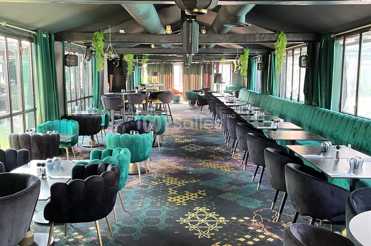 Salle de restaurant lumineuse avec tables alignées, chaises en velours coloré et décoration végétale suspendue.