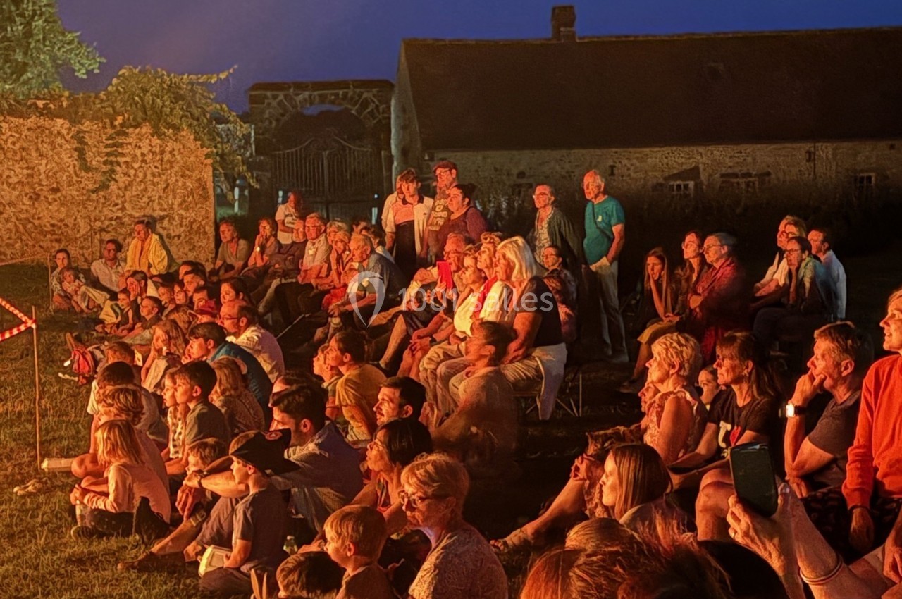 Groupe de personnes assises en plein air, attentives à une scène illuminée, devant un bâtiment en pierre au crépuscule.