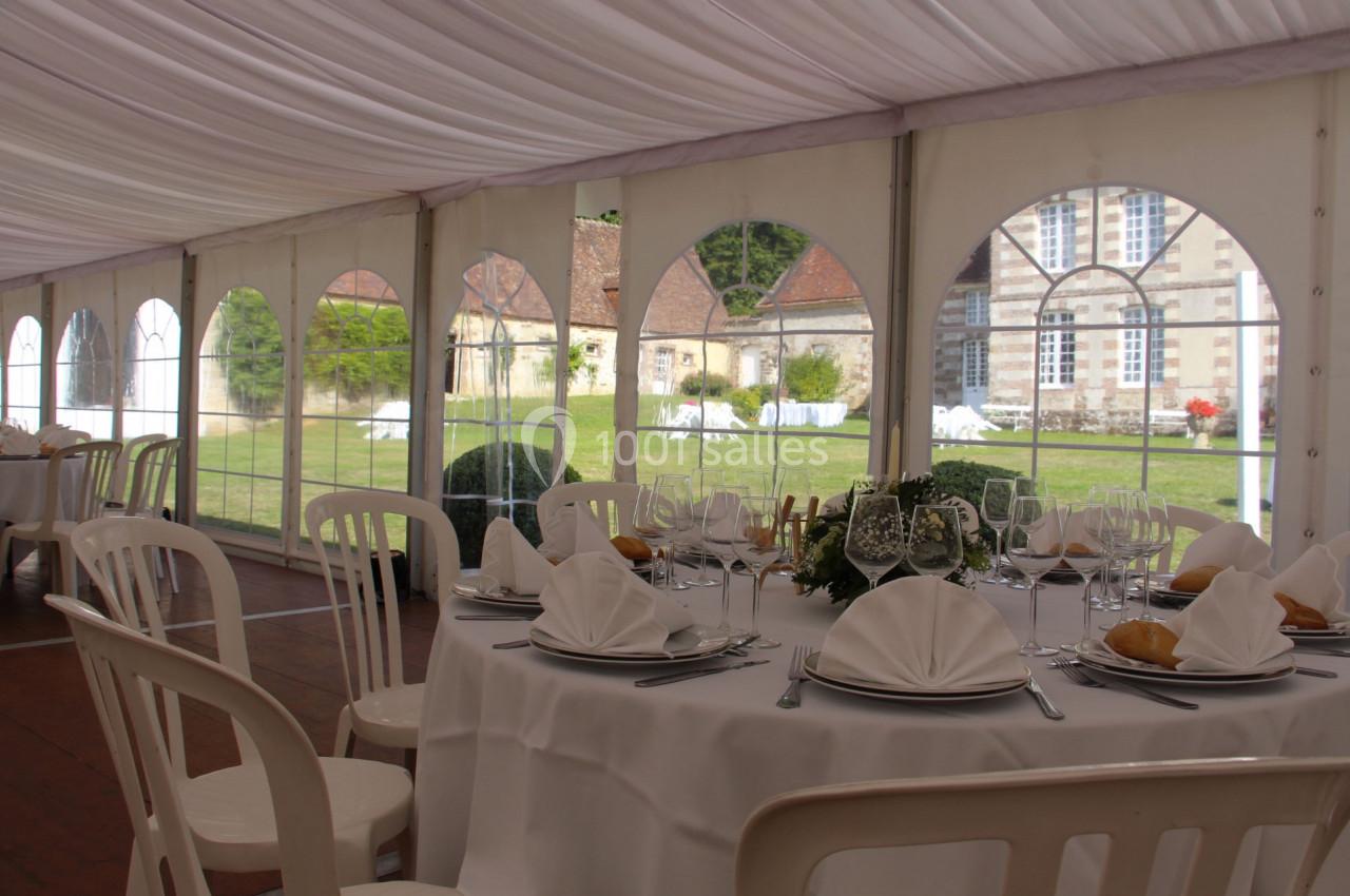 Salle de réception sous une tente avec tables dressées, nappes blanches et vue sur un jardin et un bâtiment ancien.