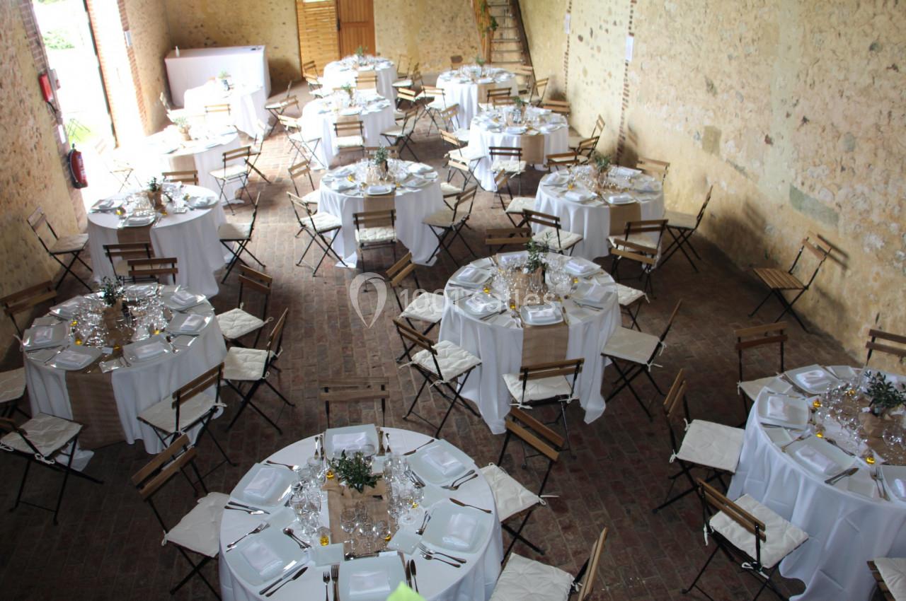 Salle rustique aménagée pour un événement, avec des tables rondes dressées, nappes blanches et chaises pliantes.