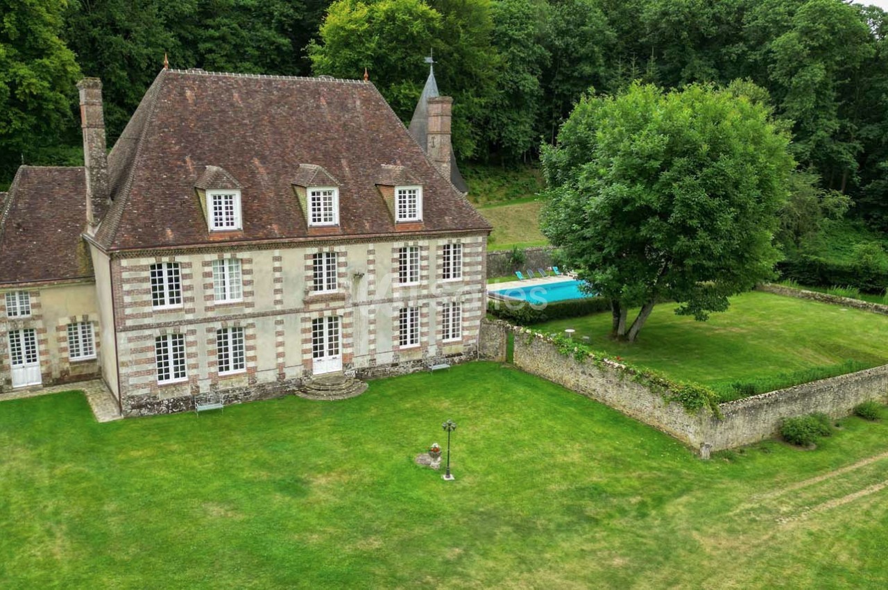 Manoir ancien en pierre avec toit en ardoise, grand jardin, piscine et forêt en arrière-plan.