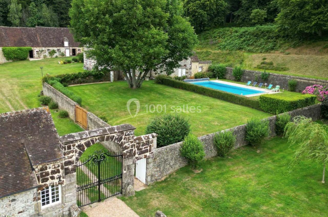 Vue aérienne d'un jardin clôturé avec piscine, pelouse bien entretenue, arbres et bâtiments en arrière-plan.