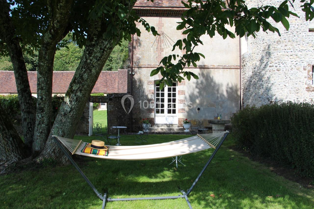 Hamac beige installé sous un arbre dans un jardin, près d'une maison en pierre avec des murs partiellement en briques.
