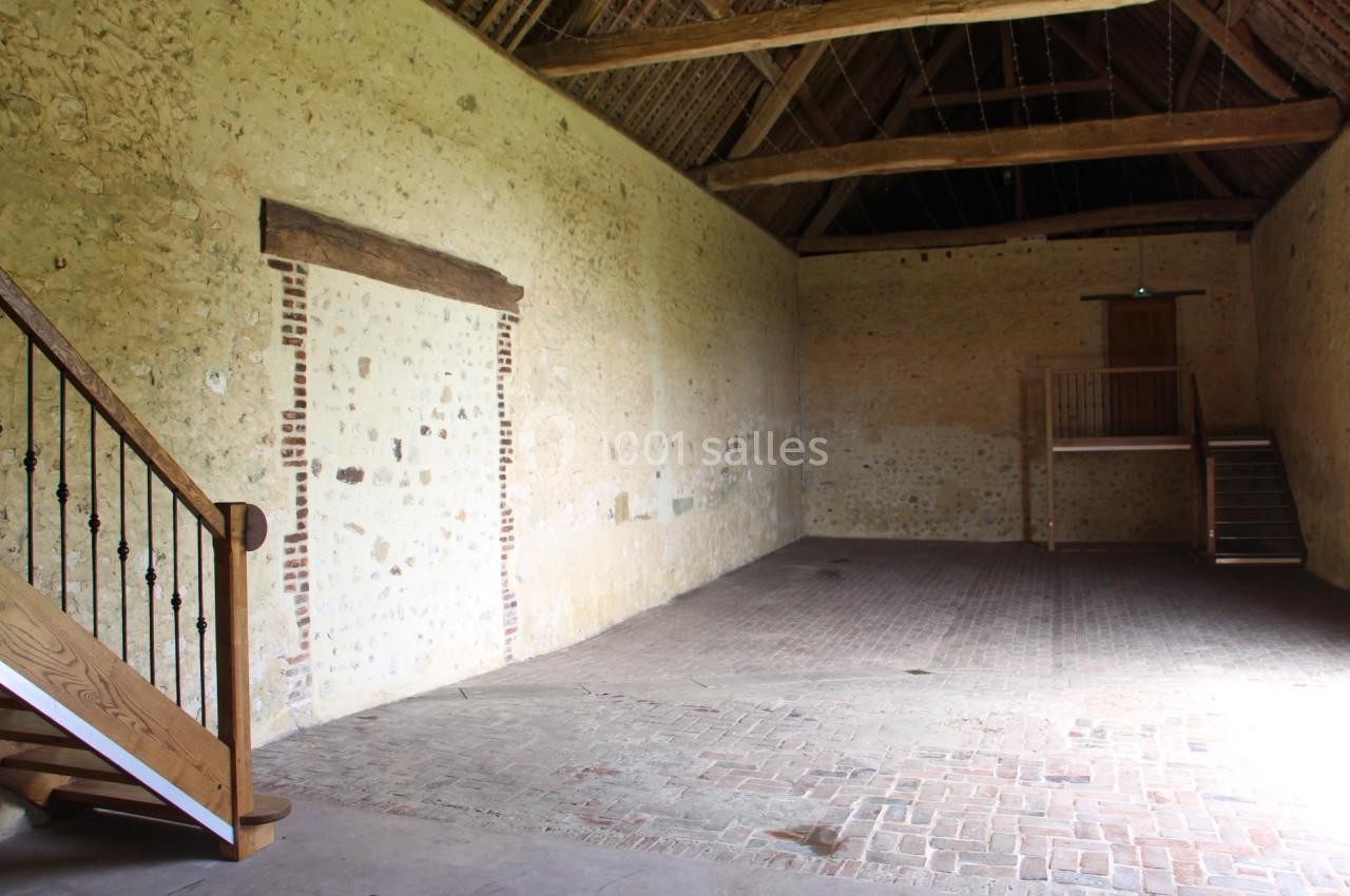 Intérieur d'une grange rénovée avec murs en pierre, sol en briques, escalier en bois et mezzanine.