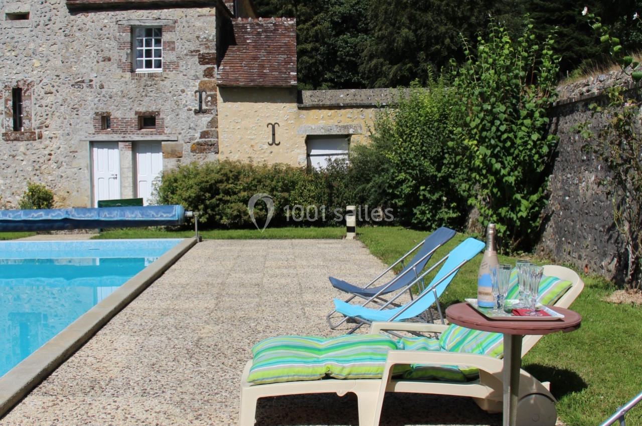 Chaises longues et table basse près d'une piscine, avec un bâtiment en pierre et des arbres en arrière-plan.