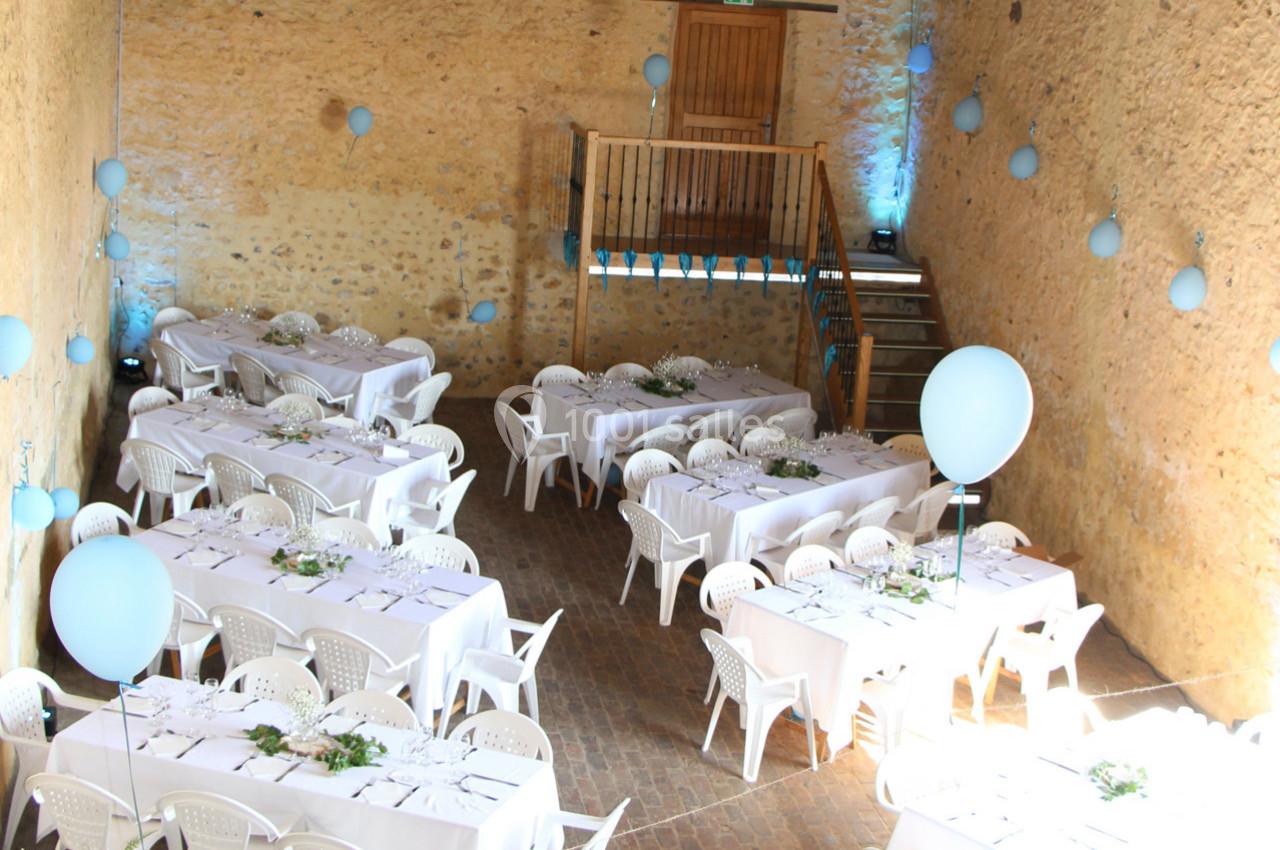 Salle en pierre décorée de ballons bleus, aménagée avec des tables blanches dressées pour un événement.