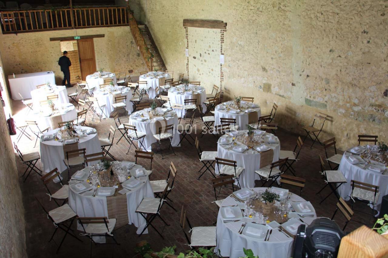 Salle de réception rustique avec des tables rondes dressées, nappes blanches et chaises en bois pliantes.