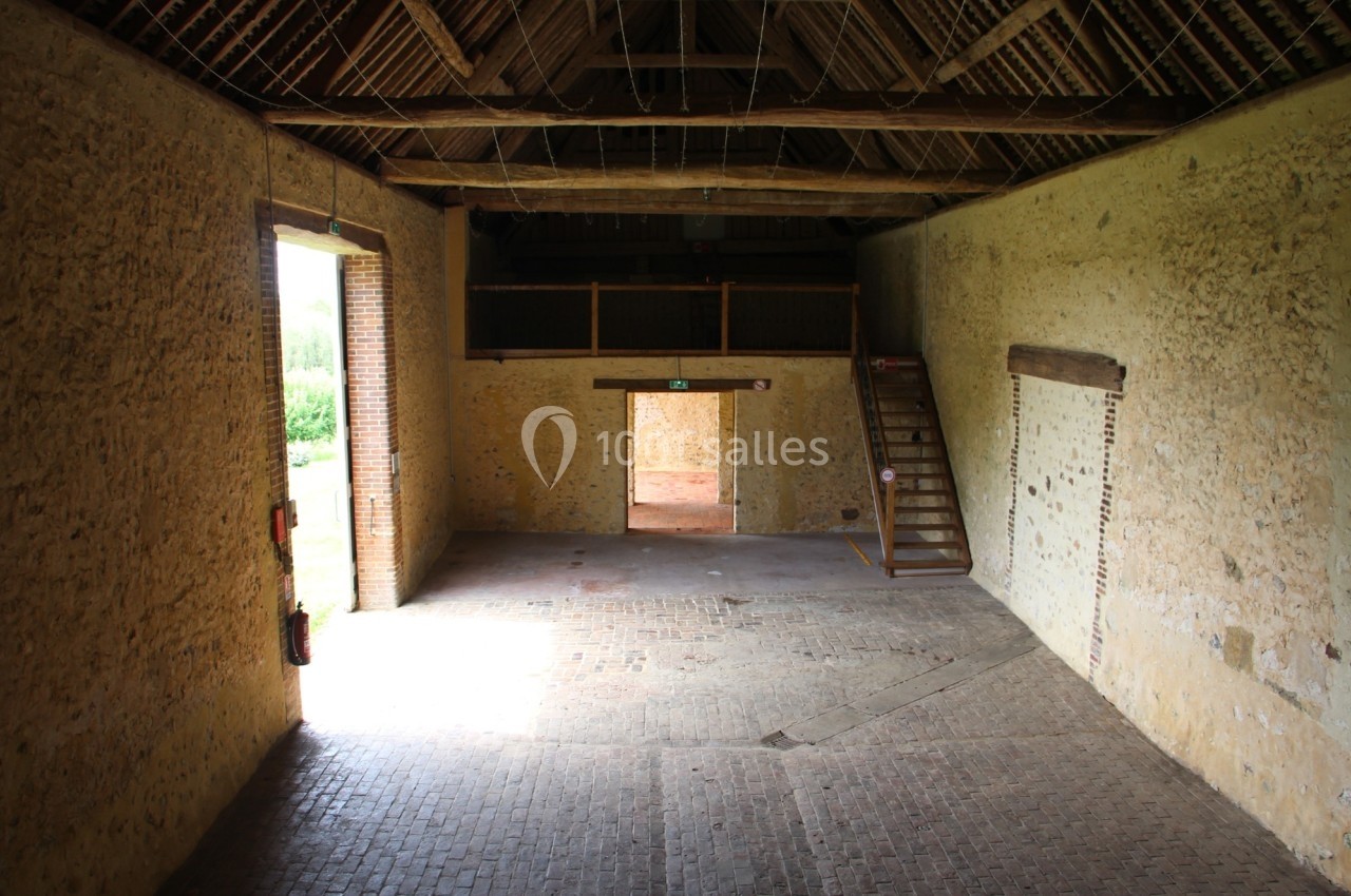 Intérieur d'une grange en pierre avec un sol en briques, une grande porte ouverte et un escalier en bois sur le côté.