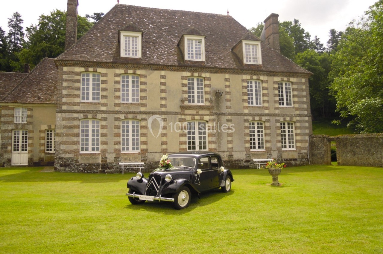 Voiture ancienne noire stationnée devant une grande maison en pierre avec jardin et arbres en arrière-plan.