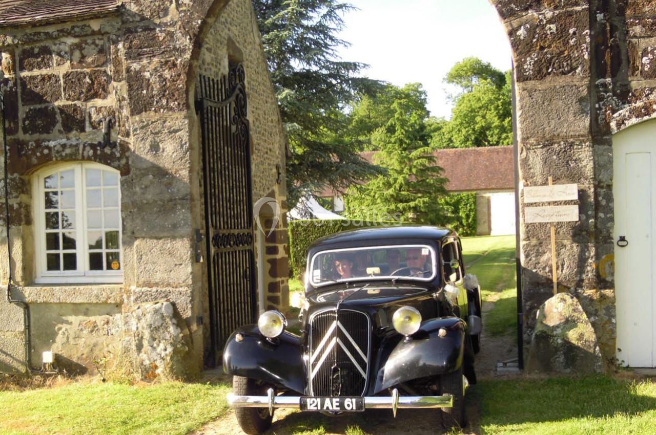 Voiture ancienne noire passant sous une arche en pierre dans un cadre rural verdoyant.