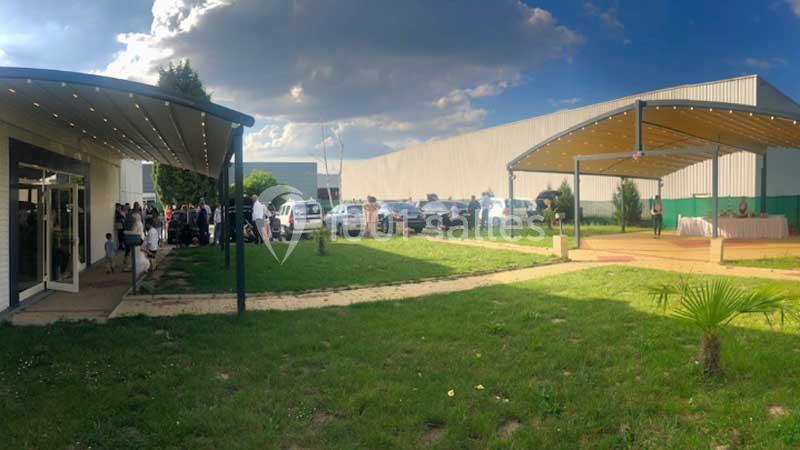 Vue d'un espace extérieur avec pelouse, pergolas, bâtiments industriels et personnes rassemblées près d'une entrée.