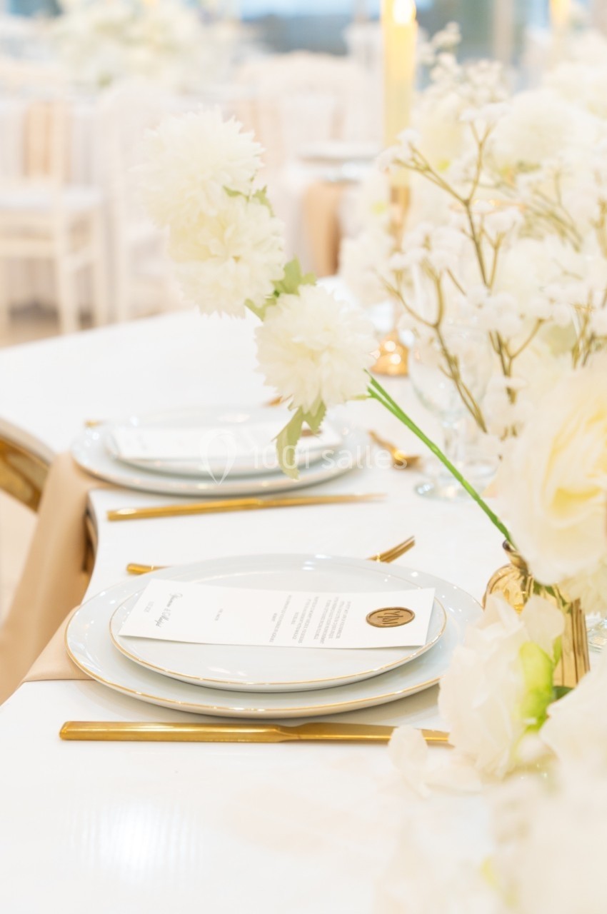 Table élégamment dressée avec vaisselle blanche, couverts dorés et décoration florale blanche.